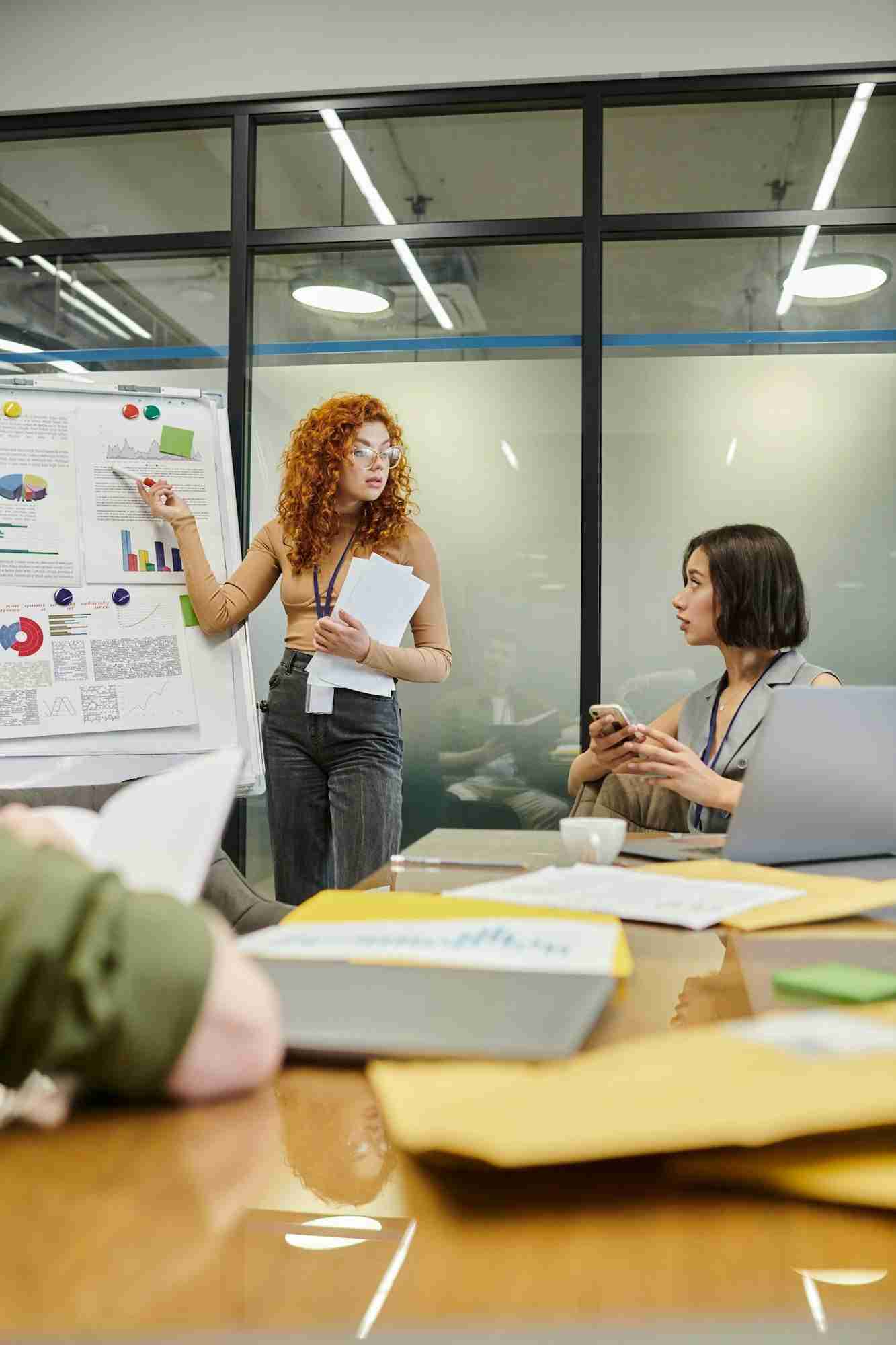 redhead businesswoman pointing at infographics on flip chart near colleagues in meeting room